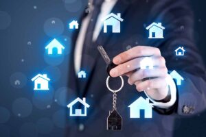Businessman In Suit Holding Keys With House Graphics Around And Dark Background.