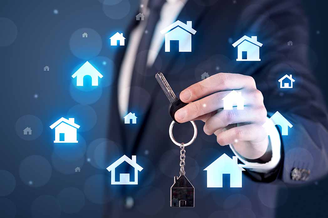 Businessman In Suit Holding Keys With House Graphics Around And Dark Background.