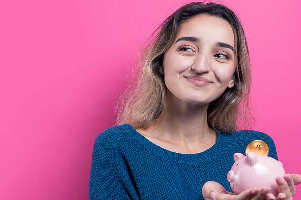 The girl puts physical Bitcoin into the piggy bank. Young girl over pink background holding a piggy bank and rejoicing, gesturing.