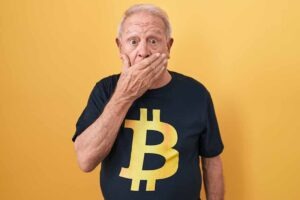 A senior man with grey hair wearing a Bitcoin t-shirt is shocked, covering his mouth with his hands in shock at the mistake. secret concept.