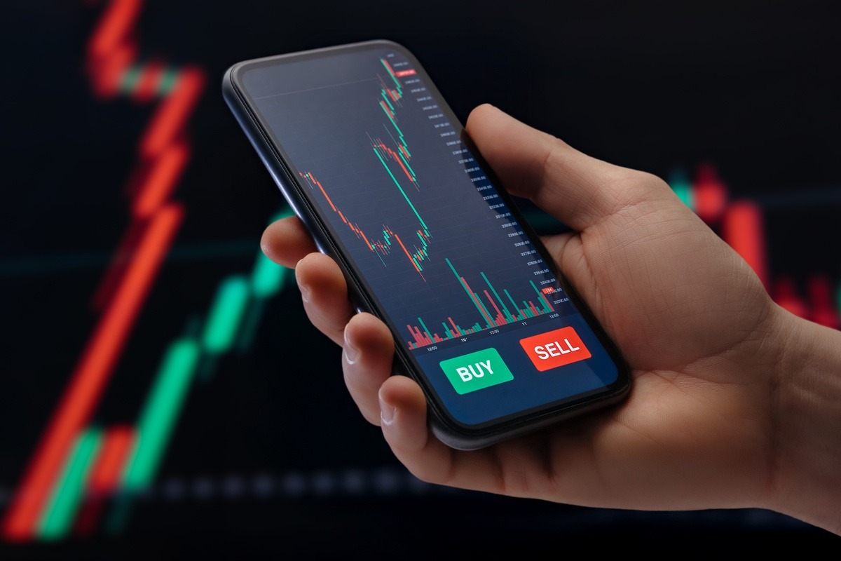 Close up, trader holding smartphone with a mobile trading platform application on screen, with candlestick chart and financial graphs for stock or crypto investments on background.